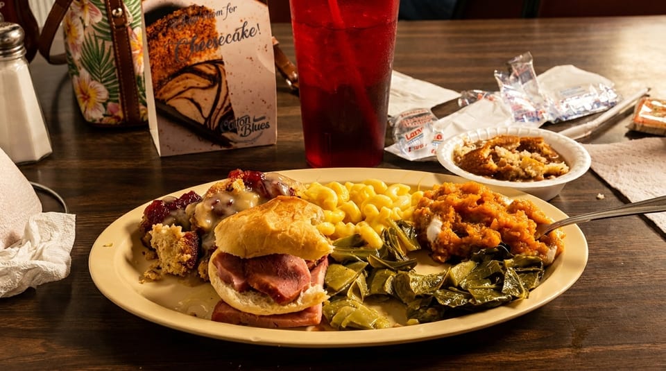A plate of Southern comfort food with ham biscuit, mac and cheese, collard greens, and sweet potatoes.