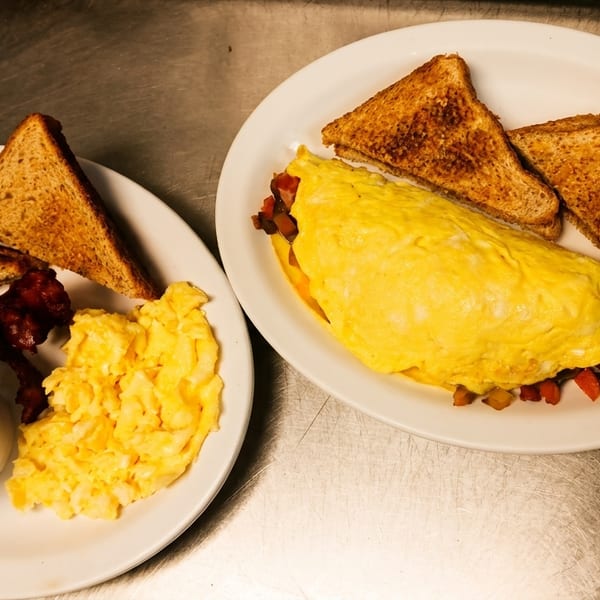 Two breakfast plates with omelets, scrambled eggs, bacon, grits, and toast on a metal surface.