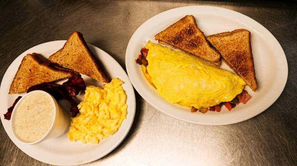 Two breakfast plates with omelets, scrambled eggs, bacon, grits, and toast on a metal surface.