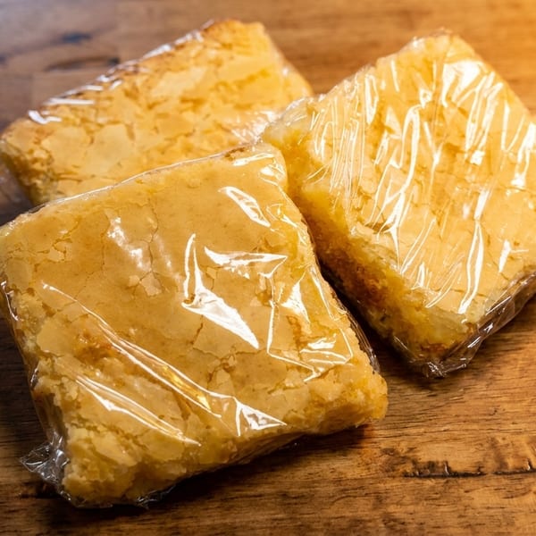 Three individually plastic-wrapped blondie bars with crinkled tops sit on a rustic wooden surface.